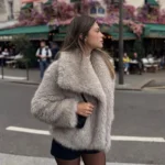 A woman wearing a stylish faux fur coat walking on city street with outdoor cafes and pink flowers i.