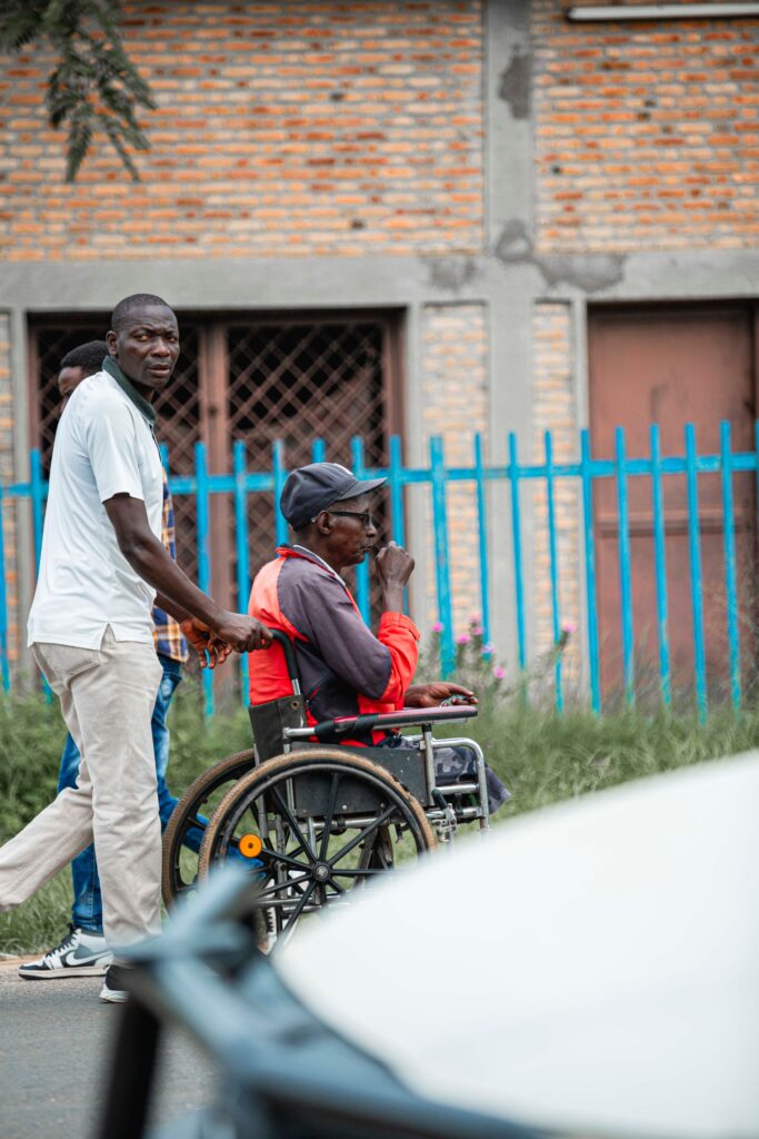 A younger man pushes an older man in a wheelchair along a quiet street beside a blue fence