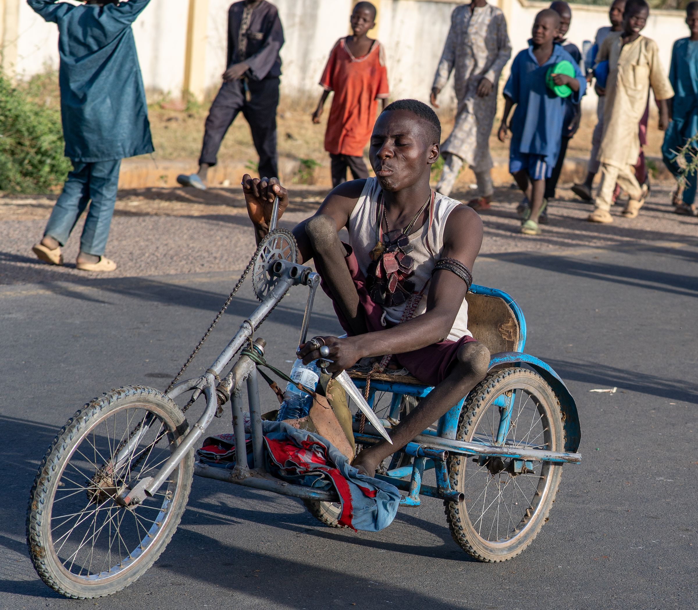 A man powers a hand-cranked tricycle wheelchair along a busy street while people walk behind him