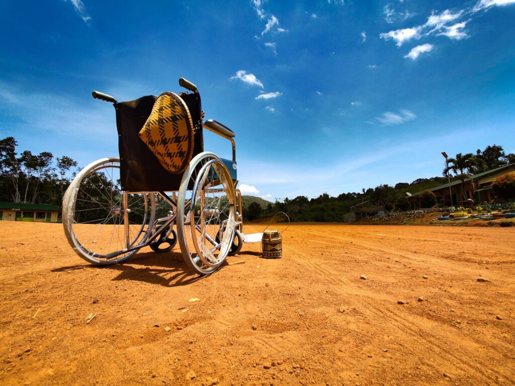 An empty wheelchair stands on a rough dirt track under a bright sky