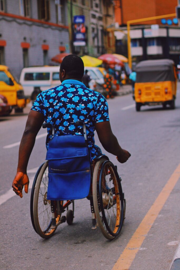 A man pushes his wheelchair along the edge of a busy city road with traffic behind him