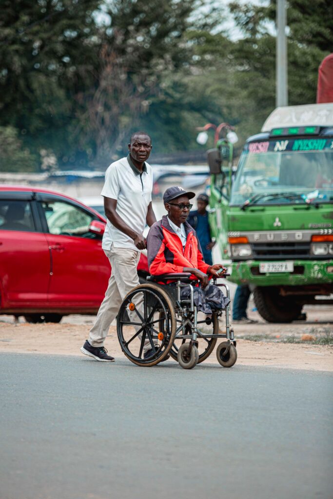 A man walks beside a woman in a wheelchair on a street next to a parked truck