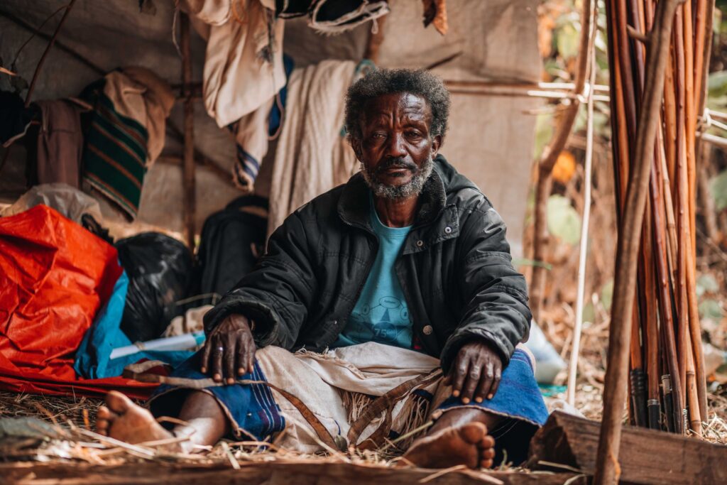 An older man sits on the ground inside a makeshift shelter of sticks and tarpaulin