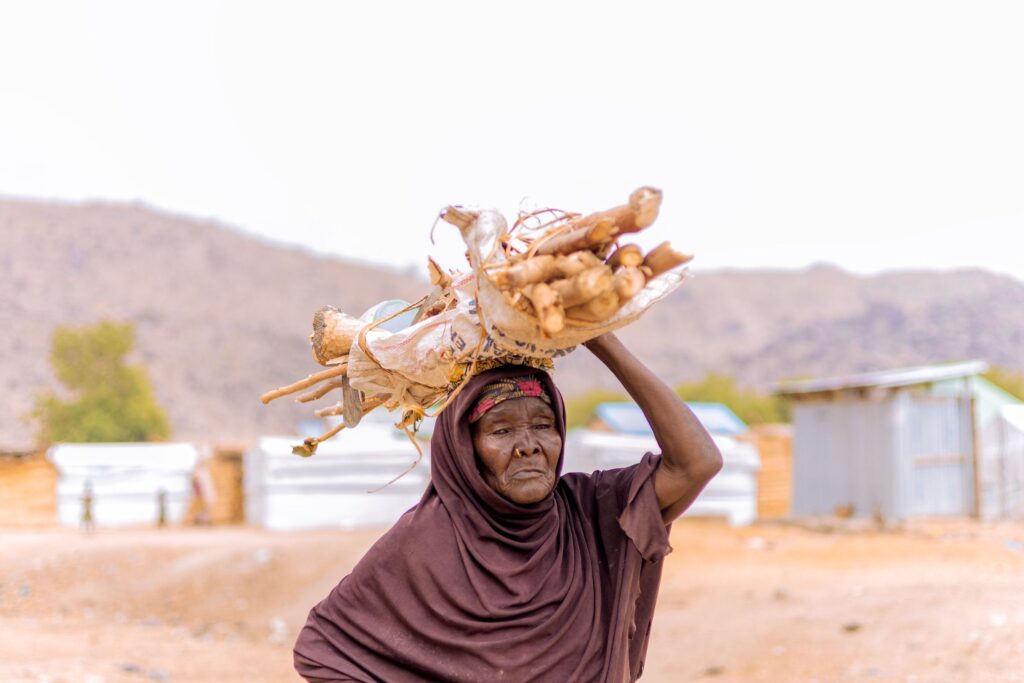 An older woman carries a bundle of firewood on her head past tarpaulin shelters in a camp