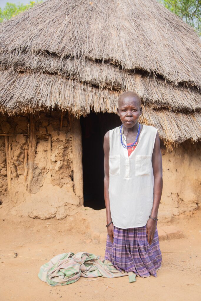 An older woman stands at the doorway of a mud-walled home with a thatched roof, a bag at her feet