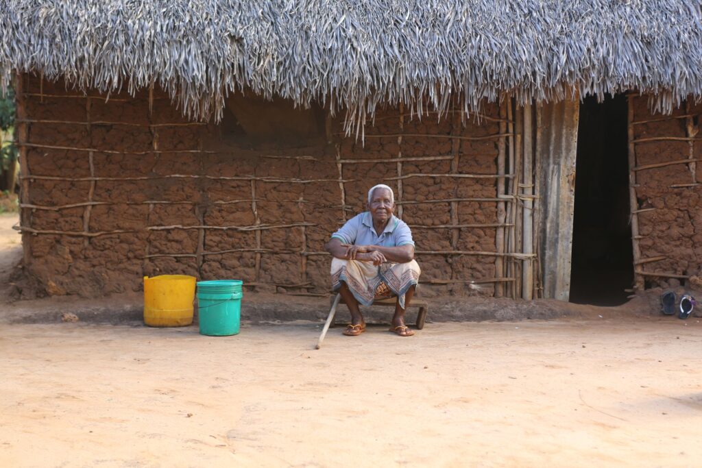 An older woman sits with a walking stick in front of a mud-walled, thatched home with water containers beside her